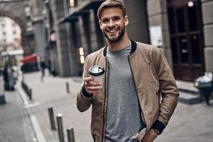 profiter du café à l'extérieur. beau jeune homme en tenue décontractée tenant une tasse jetable et souriant en marchant dans la rue de la ville photo