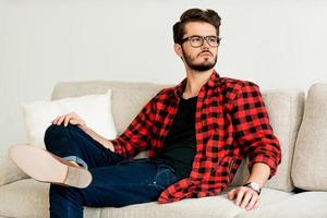 rêveur à la mode. beau jeune homme à lunettes regardant loin tout en étant assis sur un canapé photo