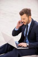 il est en contact n'importe où et n'importe quand. jeune homme confiant en tenues de soirée travaillant sur un ordinateur portable et parlant au téléphone portable assis sur le sable photo