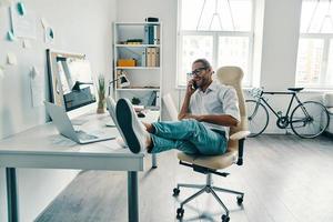 bonne conversation. beau jeune homme en chemise parlant au téléphone intelligent et souriant assis au bureau photo