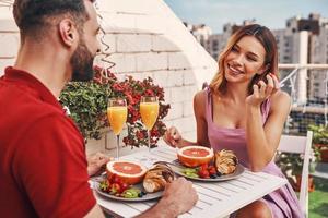 jeune couple romantique en vêtements décontractés prenant son petit déjeuner et souriant tout en étant assis sur la terrasse sur le toit à l'extérieur photo