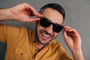 se sentir ludique. vue de dessus du beau jeune homme ajustant ses lunettes de soleil et souriant en se tenant debout sur fond gris photo