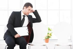 Fatigué d'attendre. jeune homme ennuyé en tenues de soirée assis à la chaise et tenant la tête dans la main tandis que d'autres chaises se tiennent dans une rangée photo