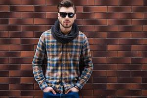 confiant et élégant. beau jeune homme appuyé contre le mur de briques et tenant les mains dans les poches en se tenant debout contre le mur de briques photo