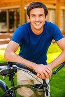 moi et mon vélo. beau jeune homme se penchant à vélo et souriant tout en se tenant à l'extérieur et contre la maison photo
