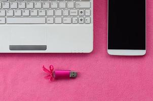 une carte mémoire flash usb rose brillante avec un arc rose se trouve sur une couverture en tissu polaire rose clair doux et poilu à côté d'un ordinateur portable et d'un smartphone blancs. conception de cadeau féminin classique pour une carte mémoire photo