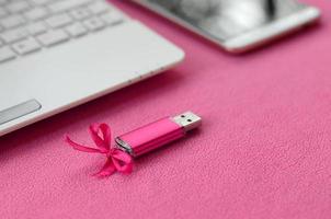 une carte mémoire flash usb rose brillante avec un arc rose se trouve sur une couverture en tissu polaire rose clair doux et poilu à côté d'un ordinateur portable et d'un smartphone blancs. conception de cadeau féminin classique pour une carte mémoire photo