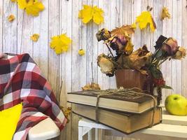 plaid à carreaux sur un fauteuil et une table en bois avec des livres vintage attachés avec de la ficelle et un vase, herbier, pomme verte sur fond de planches de bois et feuilles d'automne jaunes. photo