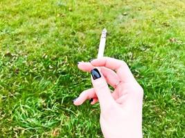 la main d'une fille qui fume, une femme avec de beaux doigts et une alliance en or et une manucure noire sur les ongles tenant une cigarette allumée sur fond d'herbe verte photo