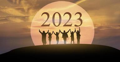 bonne année 2023, continuez à vous battre ensemble, silhouette de 2023 lettres sur la montagne avec des hommes d'affaires levés les bras dans le concept de travail d'équipe au lever du soleil. photo