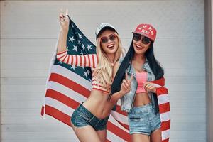 filles américaines. deux jeunes femmes espiègles gesticulant signe de paix et tenant le drapeau américain tout en se tenant contre la porte du garage photo