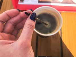 une femme tient avec une belle manucure sur ses doigts un tube dans une tasse de café naturel fort et chaud noir rapide de la restauration rapide dans un verre en papier photo