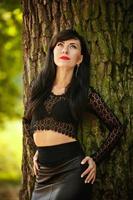 fille brune sexy dans un chemisier en dentelle et une jupe en cuir avec des talons hauts posant près d'un arbre dans les bois photo