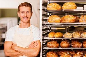 j'aime mon travail beau jeune boulanger en tablier gardant les bras croisés et souriant tout en se penchant sur le plateau avec du pain frais photo