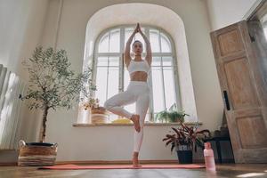 belle jeune femme regardant concentrée tout en pratiquant le yoga devant la fenêtre à la maison photo