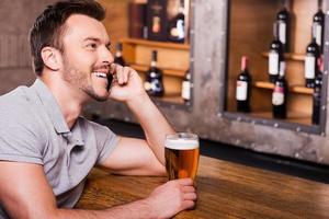 vous attend, copain vue latérale d'un jeune homme joyeux tenant un verre de bière et parlant au téléphone portable assis au comptoir du bar photo