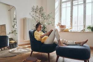 jolie jeune femme lisant un livre tout en se relaxant dans un fauteuil confortable à la maison photo