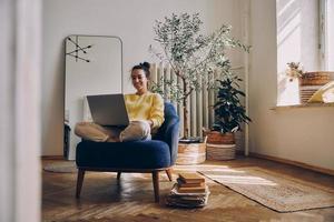 belle jeune femme utilisant un ordinateur portable et souriant assis dans une chaise confortable à la maison photo