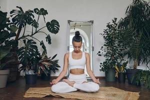 jeune femme concentrée méditant assise sur le sol avec des plantes d'intérieur tout autour d'elle photo