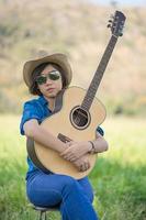 les femmes cheveux courts portent un chapeau et des lunettes de soleil s'assoient en jouant de la guitare dans le champ d'herbe photo