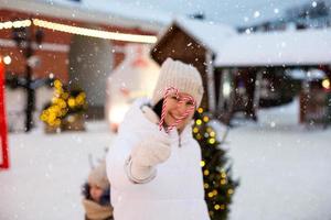 femme tient dans ses mains une canne en bonbon en forme de coeur en plein air dans des vêtements chauds blancs sur le marché festif d'hiver. guirlandes de guirlandes lumineuses décorées de ville de neige pour le nouvel an. ambiance de noël photo