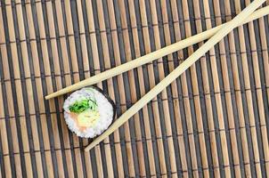 un rouleau de sushi et des baguettes en bois reposent sur un tapis de couture en paille de bambou. cuisine asiatique traditionnelle. vue de dessus. minimalisme à plat tourné avec espace de copie photo