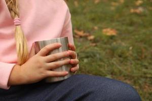 petite fille en sweat-shirt rose clair tient dans les mains une tasse en acier inoxydable avec du thé chaud. ambiance hygge d'automne. plein air, pique-nique, camping, voyage, concept de trekking. image recadrée, pas de visage, méconnaissable. photo