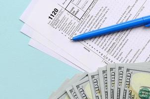 Le formulaire d'impôt 1120 se trouve près de cent billets d'un dollar et d'un stylo bleu sur fond bleu clair. déclaration de revenus des sociétés américaines photo