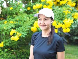 femme asiatique en bonne santé en tenue de sport noire écoutant de la musique à partir d'écouteurs sans fil, debout dans un jardin avec des fleurs jaunes, souriant et regardant ailleurs. photo