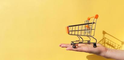 sur la main d'une femme se trouve un petit chariot en métal vide d'un supermarché. l'objet est sur fond jaune. photographie horizontale. notion de commerce. acheteur. notion d'achat. fermer. espace de copie. photo