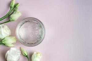 pot avec produit cosmétique transparent, gel ou crème sur fond rose avec des fleurs. emballage sans marque avec crème antioxydante pour les soins personnels. vue d'en-haut. photo