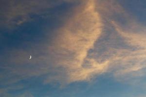 une lune croissante dans le ciel bleu parmi les nuages éclairés par le soleil au coucher du soleil photo