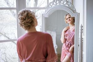 une femme heureuse regarde son reflet dans un miroir en robe de soie rose près de la fenêtre photo