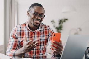 homme afro-américain à lunettes parlant en ligne par appel vidéo, tenant un smartphone, utilisant des applications mobiles photo