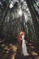 heureux couple élégant jeunes mariés dans la forêt verte le jour de l'été. la mariée en longue robe blanche et le marié en costume rouge s'étreignent. Jour de mariage. photo