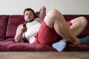 l'homme tient une télécommande de télévision allongée sur le canapé, il a trouvé un film intéressant et est agréablement surpris. concept de style de vie paresseux. photo