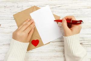 main de fille écrivant une lettre d'amour le jour de la saint-valentin. carte postale faite à la main. une femme écrit sur une carte postale pour la célébration des vacances du 14 février. mise à plat. vue de dessus photo
