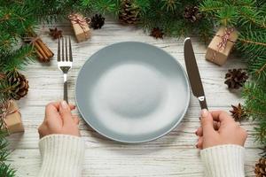 vue en perspective fille tient une fourchette et un couteau à la main et est prête à manger. assiette vide en céramique ronde sur fond de noël en bois. concept de plat de dîner de vacances avec un décor de nouvel an photo