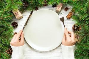 vue de dessus la fille tient une fourchette et un couteau à la main et est prête à manger. assiette vide en céramique ronde sur fond de noël en bois. concept de plat de dîner de vacances avec un décor de nouvel an photo