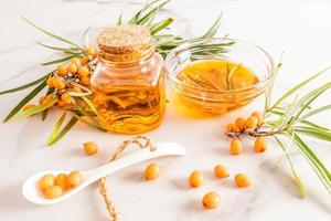 huile d'argousier naturelle dans une bouteille en verre avec un bouchon et un bol de baies d'argousier sur fond de marbre blanc. un remède d'origine végétale. photo