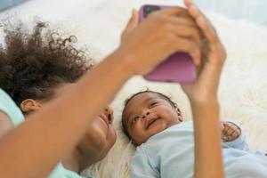 jeune mère afro-américaine et nouveau-né faisant un appel vidéo et souriant saluer. bonne sœur aînée prenant selfie avec petit frère bébé allongé sur le lit en regardant un téléphone portable souriant sans souci. photo