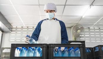 jeune homme travailleur ou inspecteur de la qualité en vêtements de travail et avec un masque de protection sur son visage travaillant à vérifier l'eau potable en bouteille dans l'usine d'eau potable avant expédition. entreprise d'eau potable photo