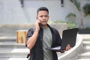 jeune homme asiatique séduisant utilisant un ordinateur portable dans un espace de travail partagé avec un visage heureux et appelant son amie photo