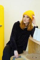 une jeune femme aux cheveux blonds dans un chapeau de tricot jaune est assise dans un café près d'un réfrigérateur jaune et regarde la caméra. photo