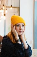 une jeune femme aux yeux bleus et aux cheveux blonds dans un bonnet jaune est assise dans un café et regarde par la fenêtre. photo