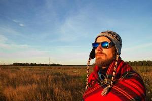 jeune homme heureux dans le bonnet tricoté, les lunettes de soleil et la couverture se réunit du lever du soleil sur le fond d'un champ et d'un ciel bleu. photo