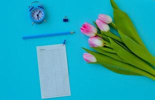 bureau à domicile plat pour femme. espace de travail avec bouquet de tulipes roses, réveil, crayon et un morceau de papier pour les notes sur fond bleu. fond féminin vue de dessus. photo