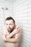 homme nu se serre dans la douche, il s'est figé et frissonne, car tout à coup l'eau chaude a disparu et il ne restait que de l'eau froide. photo