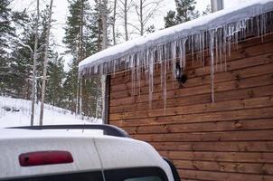 des glaçons pendent du toit d'une maison en rondins près de laquelle une voiture est garée négligemment, qui peut être endommagée par la chute de la banquise. photo