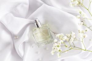 une bouteille transparente de produit cosmétique repose sur un tissu texturé satiné blanc et de délicates fleurs de gypsophile. un modèle pour le parfum.
